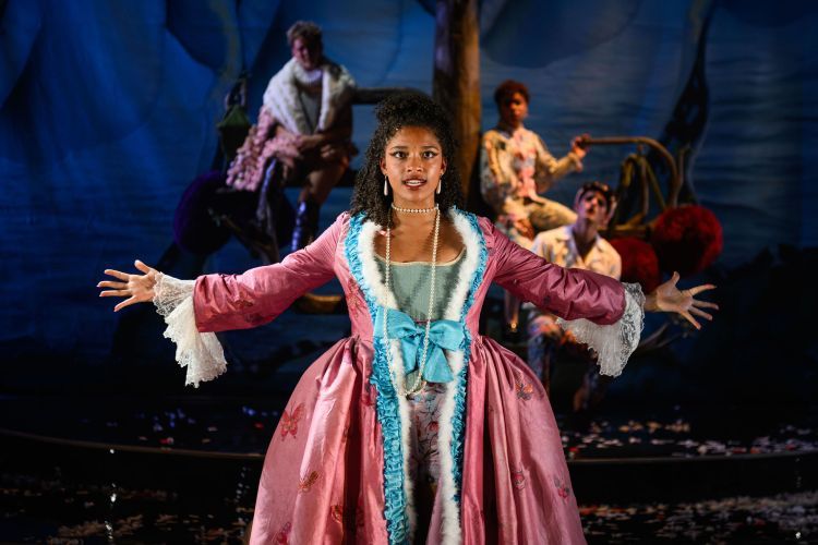Scene from Orlando - actor in blue and pink 16th-century dress standing with arms stretched out to sides